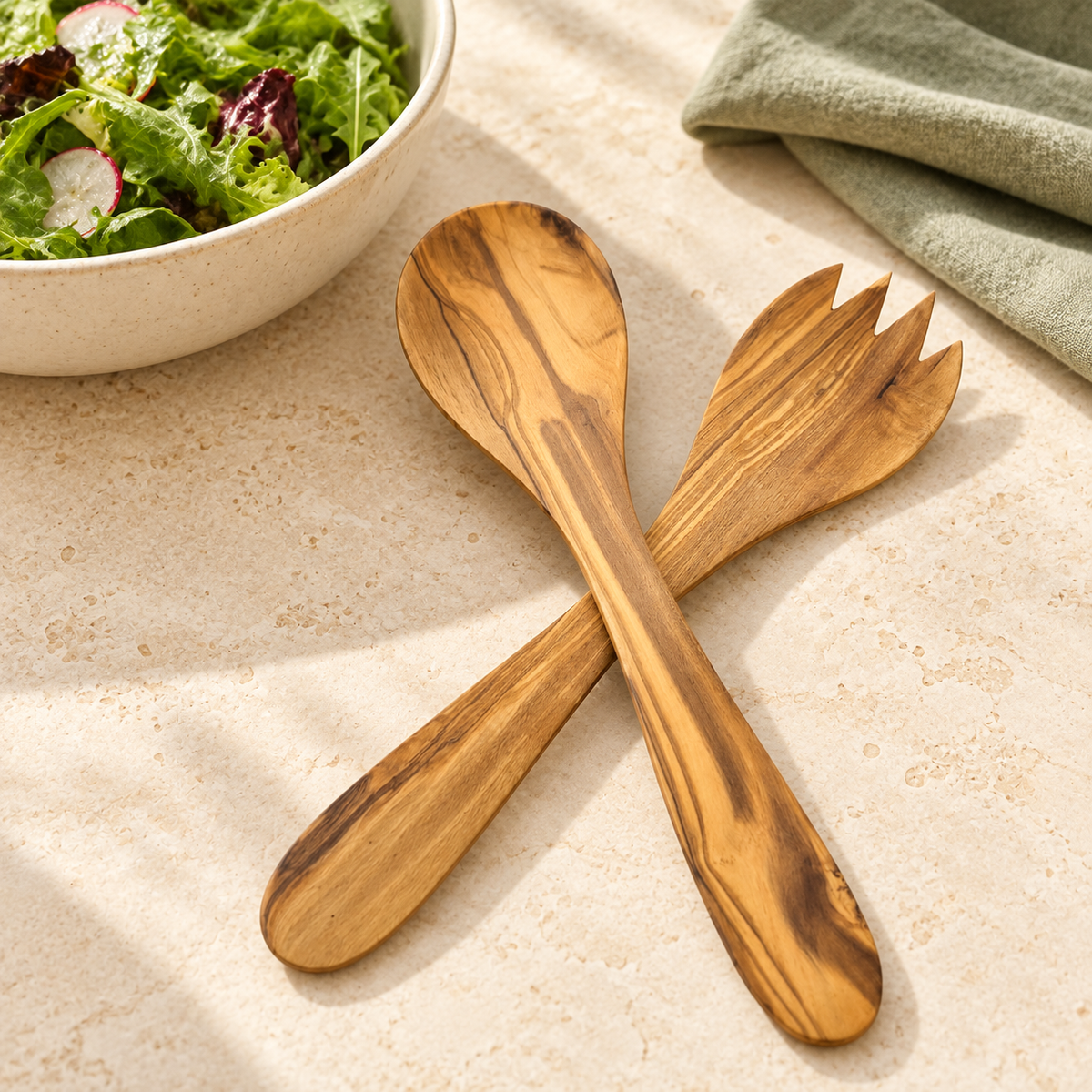 Zweiteiliges Olivenholz-Salatbesteck neben einer Salatschale auf hellem Tisch; Schale, Salat und Leinen sind Dekoration